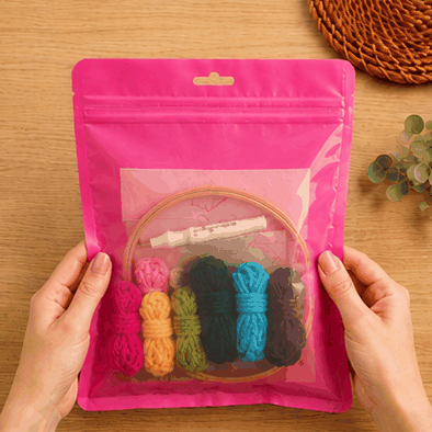 Hands holding a pink resealable pouch with an embroidery hoop, yarn bundles, and a pen inside.
